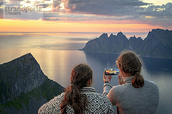 Zwei junge skandinavische Frauen fotografieren den Fjord bei Sonnenuntergang mit ihrem Smartphone  Insel Senja  Provinz Troms  Norwegen  Skandinavien  Europa