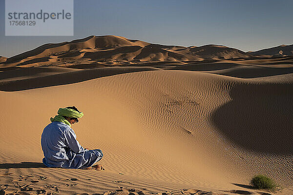 Ein Berber in traditioneller Kleidung sitzt inmitten der Sanddünen von Erg Chebbi  Sahara-Wüste  Marokko  Nordafrika  Afrika