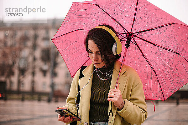 Junge Frau benutzt Smartphone und hält Regenschirm in der Stadt