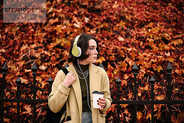 Junge Frau hört Musik über Kopfhörer und steht im Herbst in der Stadt