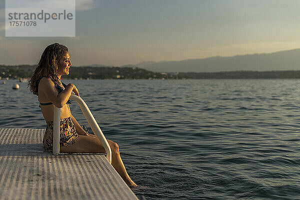Glückliches lateinamerikanisches Mädchen  das an einem warmen Nachmittag auf einem Steg am See sitzt