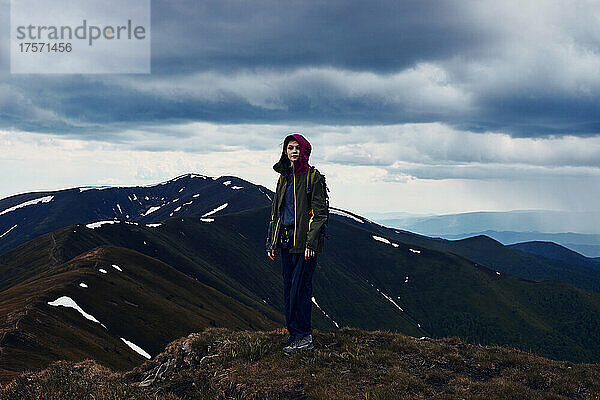 Junge Bergwandererin auf einem Berggipfel vor bewölktem Himmel.