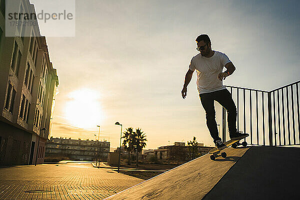 Erwachsener Mann geht bei Sonnenuntergang mit einem Surf-Skate die Rampe hinunter