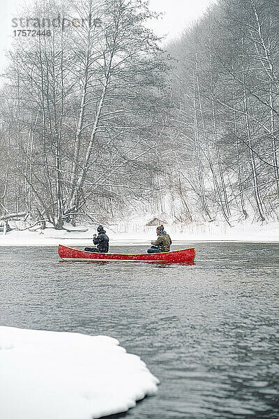 Zwei Personen paddeln in einem roten Holzkanu auf dem verschneiten Fluss.