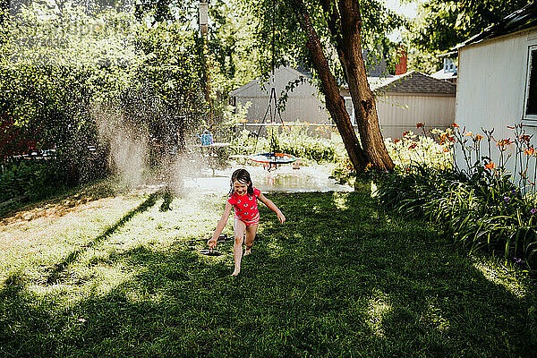 Junges Mädchen läuft im Sommer durch Sprinkleranlage im Hinterhof