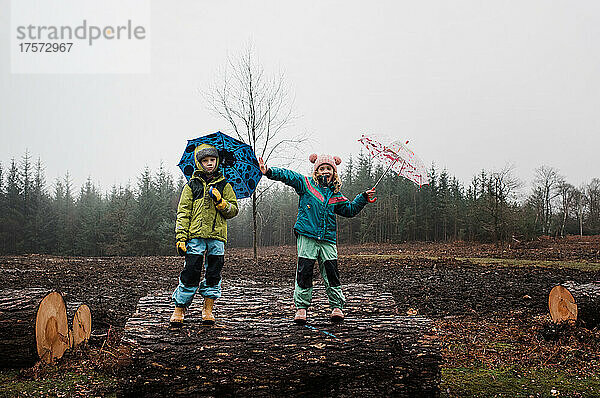 Kinder spielen draußen im Regen fröhlich mit Regenschirmen auf Baumstämmen