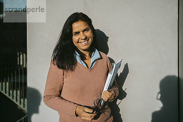 Lächelnde reife Frau mit Buch und Akte  die an einer grauen Wand in der Universität an einem sonnigen Tag steht