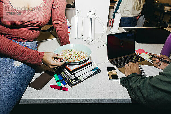 Midsection Frau mit Nudeln  während männlicher Freund mit Laptop im Studentenwohnheim