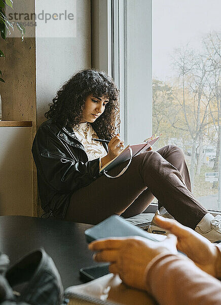 Junge Frau schreibt in ein Buch  während sie in der Universität am Fenster sitzt