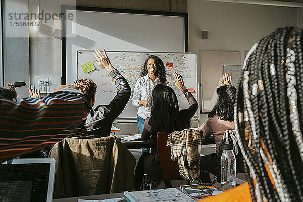 Lächelnder Professor  der Universitätsstudenten ansieht  die mit erhobenen Händen im Klassenzimmer sitzen