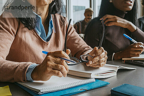 Mittelteil einer reifen Studentin  die eine Brille hält  während sie am Schreibtisch im Klassenzimmer sitzt