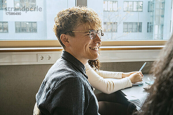 Seitenansicht eines lächelnden jungen blonden Mannes mit Brille  der im Klassenzimmer sitzt