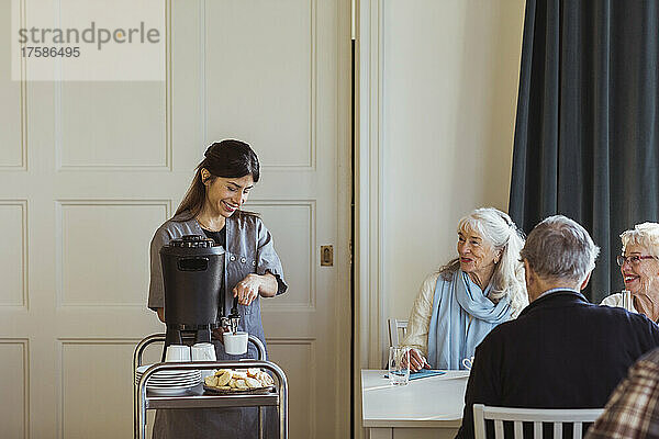 Lächelnde Mitarbeiterin im Gesundheitswesen serviert Kaffee an ältere Frauen und Männer im Altersheim