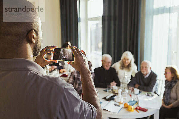 Männlicher Pfleger fotografiert ältere Männer und Frauen mit dem Smartphone im Pflegeheim
