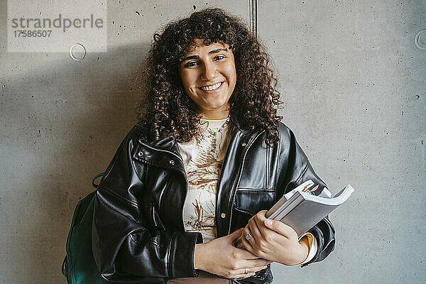 Porträt einer lächelnden jungen Frau mit Buch und Rucksack an einer grauen Wand lehnend