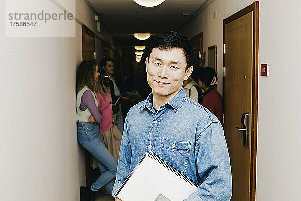 Selbstbewusster junger Mann mit Buch  während im Hintergrund rassisch gemischte Freunde auf dem Korridor eines Wohnheims zu sehen sind