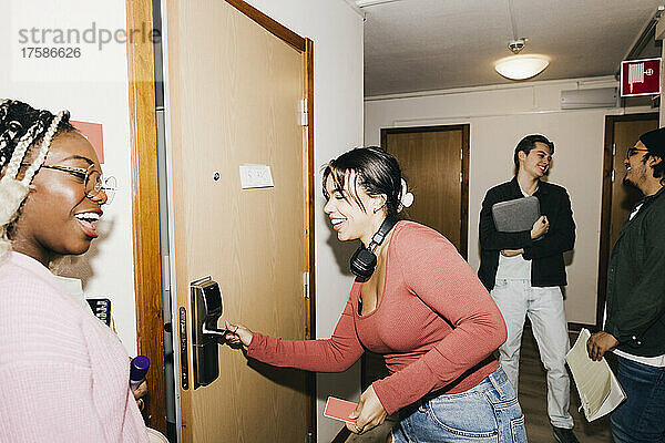 Glückliche junge Frau  die die Tür öffnet  während ihre multirassischen Freunde auf dem Flur des Studentenwohnheims stehen