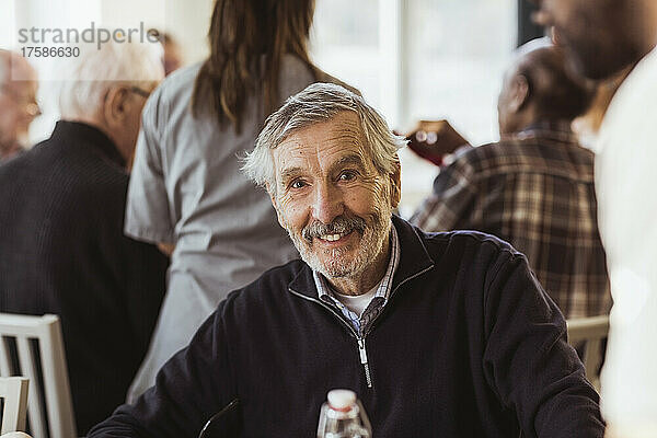 Porträt eines lächelnden älteren Mannes mit Pfleger und Freunden im Hintergrund in einem Pflegeheim