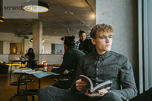 Nachdenklicher junger blonder Mann sitzt mit einem Buch in der Universitätscafeteria