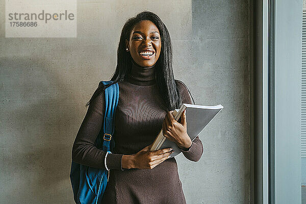 Porträt einer fröhlichen jungen Frau  die ein Buch hält und mit einem Rucksack vor einer grauen Wand steht