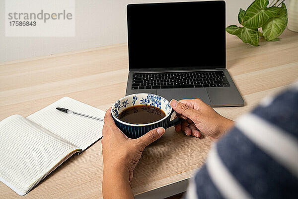 Nahaufnahme der Hände einer Frau mit Kaffeetasse am Schreibtisch  Laptop und Notizblock