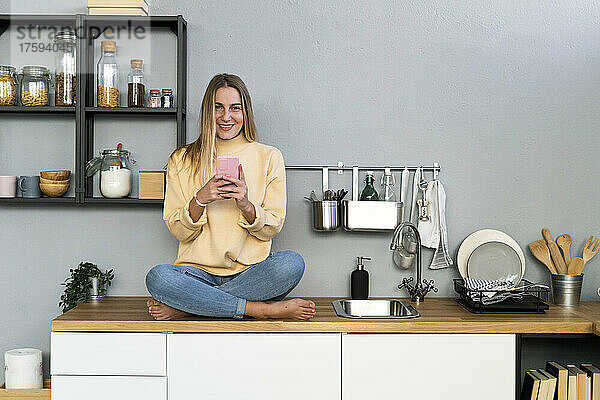 Lächelnde Frau mit Mobiltelefon sitzt zu Hause auf der Küchentheke