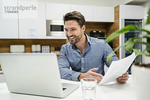Lächelnder Geschäftsmann mit Dokument  der im Büro am Laptop arbeitet