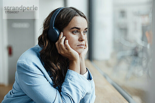 Nachdenkliche Geschäftsfrau mit Kopf in der Hand  die im Büro Musik über drahtlose Kopfhörer hört