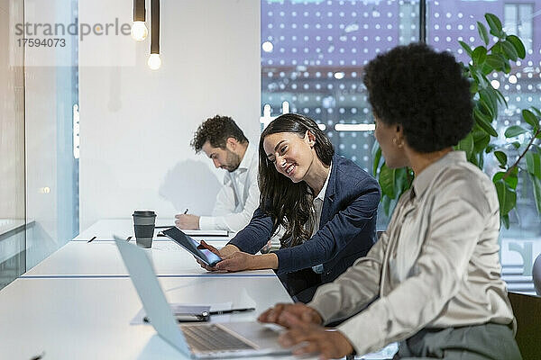 Lächelnde Geschäftsfrau teilt Tablet-PC mit Kollege am Schreibtisch im Büro