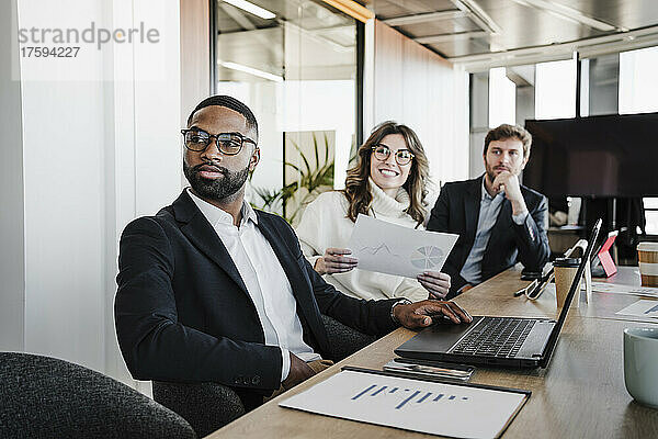 Junger Geschäftsmann mit Brille sitzt neben Kollegen am Konferenztisch im Sitzungssaal