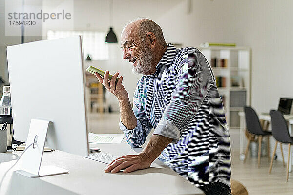 Lächelnder älterer Geschäftsmann  der über den Lautsprecher am Schreibtisch mit dem Smartphone spricht