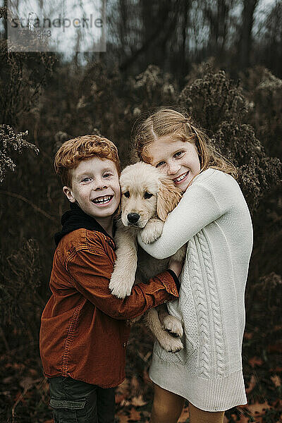 Glückliche Geschwister umarmen Golden Retriever Welpen im Wald