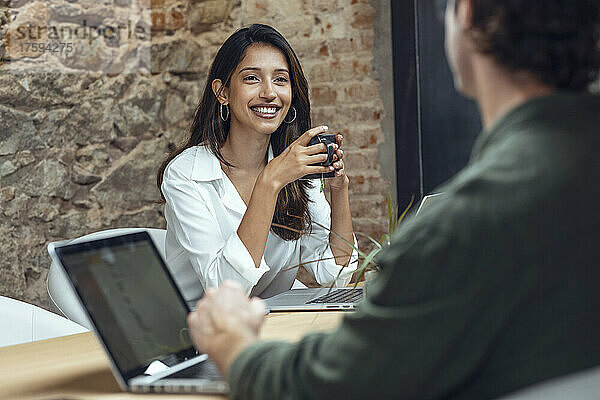 Lächelnde Geschäftsfrau hält Kaffeetasse in der Hand und diskutiert mit Geschäftsmann am Schreibtisch im Büro