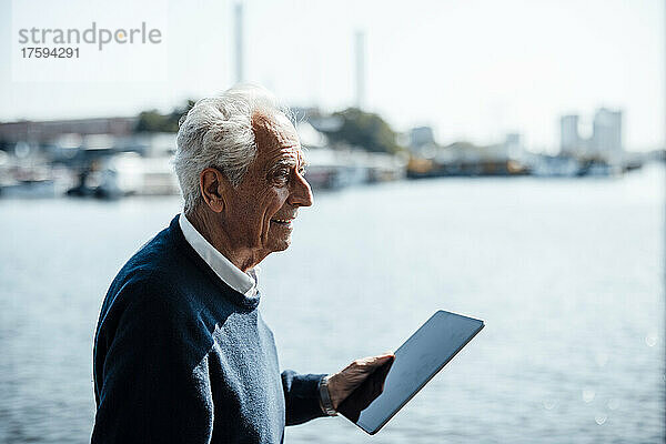 Lächelnder älterer Mann mit weißem Haar hält Tablet-PC an einem sonnigen Tag am Wasser