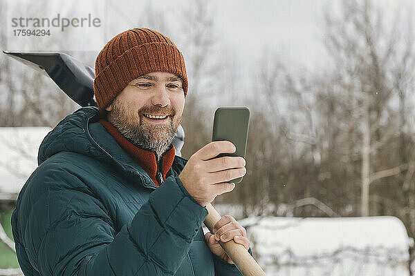 Glücklicher Mann mit Schneeschaufel und Smartphone im Winter