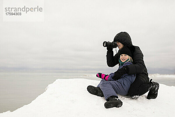 Mutter fotografiert mit der Kamera  während sie im Winter mit ihrer Tochter im Schnee sitzt