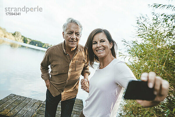 Glückliche Frau macht Selfie per Handy mit älterem Mann auf Steg am See