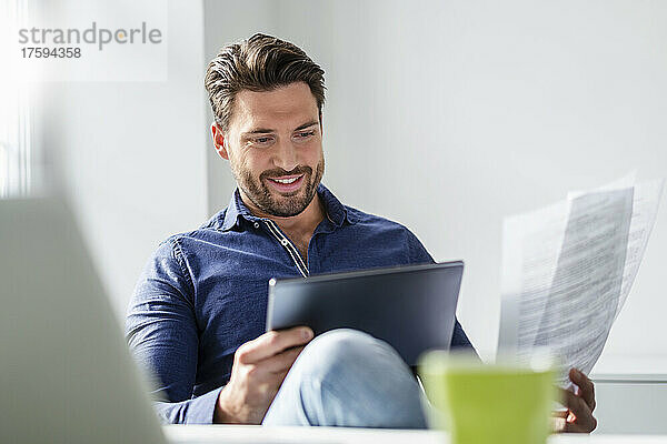 Lächelnder Geschäftsmann mit Dokument  der im Büro am Tablet-PC arbeitet