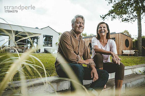 Fröhliche Frau lacht über älteren Mann  der auf Stufen im Hinterhof sitzt