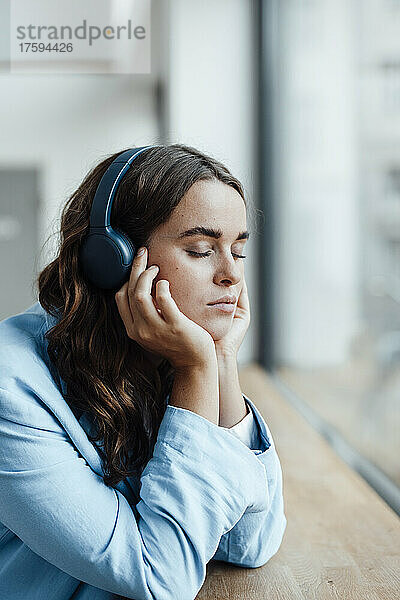 Junge Geschäftsfrau mit Kopf in den Händen  die am Schreibtisch Musik über kabellose Kopfhörer hört