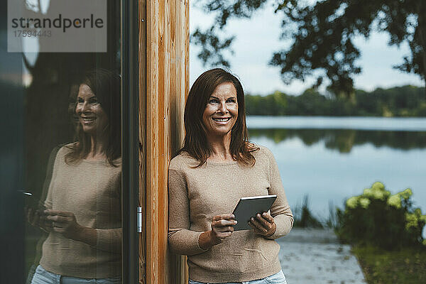 Glückliche Frau mit Tablet-PC steht an der Glaswand im Hinterhof