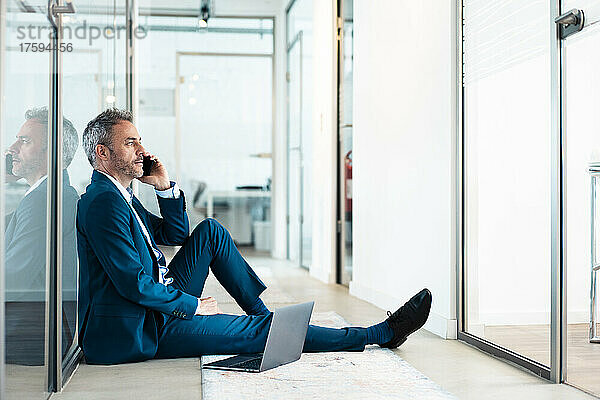 Nachdenklicher Geschäftsmann sitzt mit Laptop und telefoniert im Büroflur
