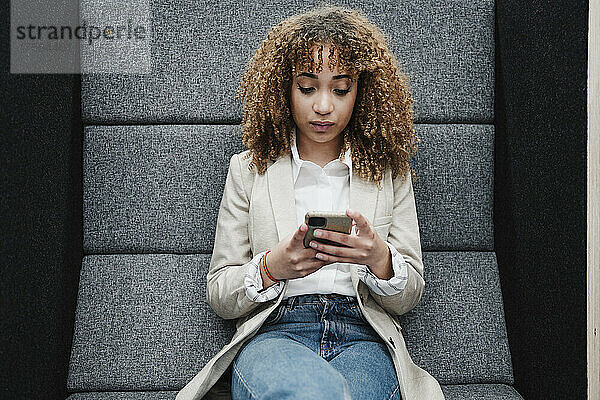 Junge Geschäftsfrau mit braunem lockigem Haar  die im Büro SMS über ihr Smartphone sendet