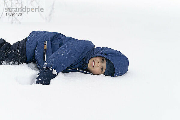 Netter Junge in Jacke  der im Winter auf Schnee liegt