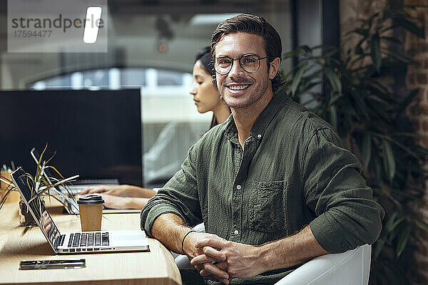 Selbstbewusster Geschäftsmann mit Laptop sitzt neben einem Kollegen am Schreibtisch im Büro