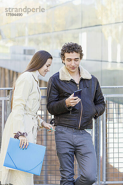 Lächelnde Frau schaut einen Freund an  der sein Smartphone auf dem Campus benutzt
