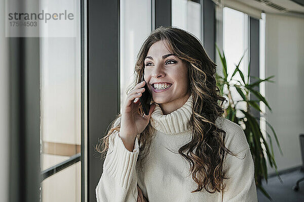 Glückliche Geschäftsfrau mit langen Haaren  die im Coworking-Büro auf dem Smartphone spricht