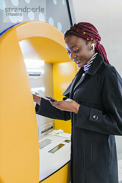 Junge Frau mit Smartphone benutzt Geldautomaten
