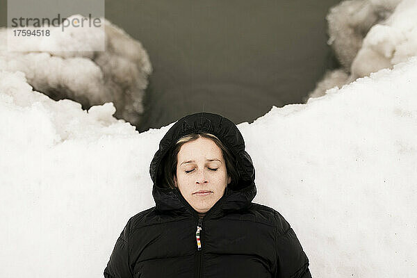Frau mit geschlossenen Augen liegt im Winter auf Schnee am See