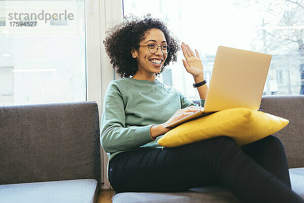 Glückliche Geschäftsfrau winkt per Videoanruf über Laptop auf dem Sofa